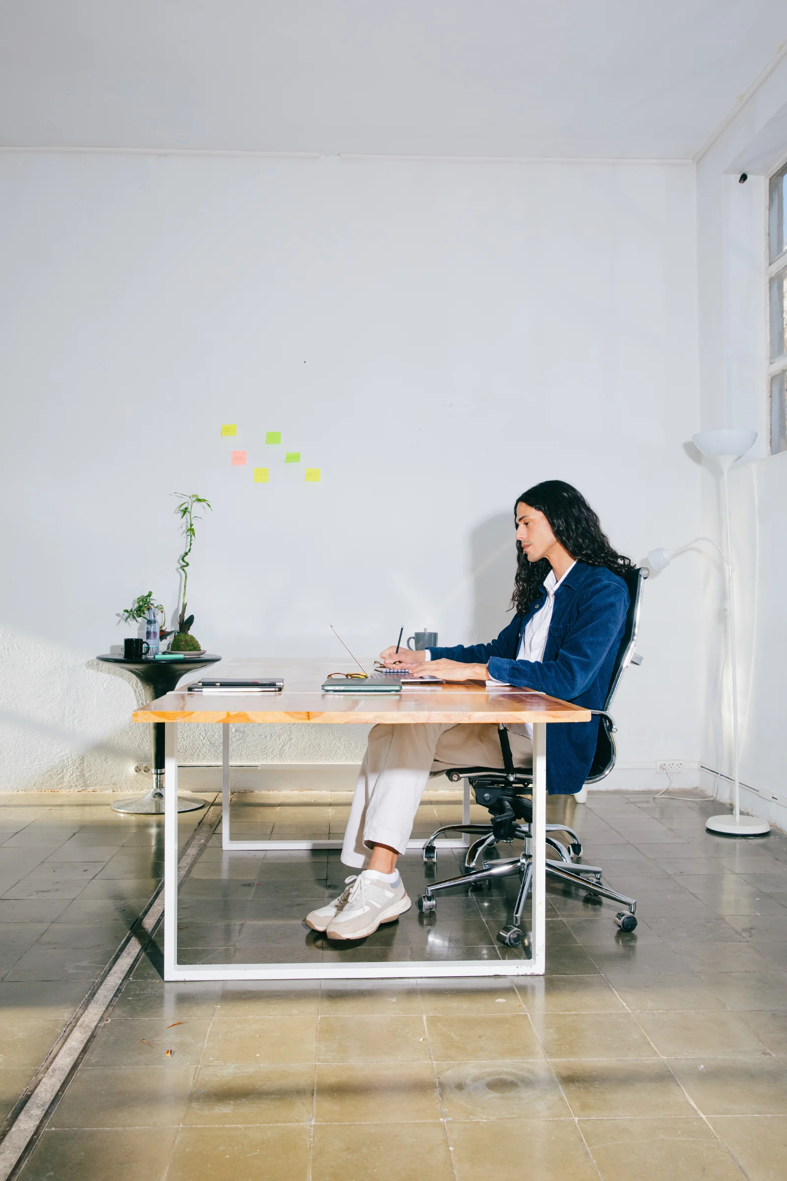 Eine Person in einem blauen Jacket und mit weißer Hose sitzt an einem Schreibtisch und arbeitet an einem Laptop. Auf dem Schreibtisch liegen Bücher, eine Brille und eine Tasse Kaffee.